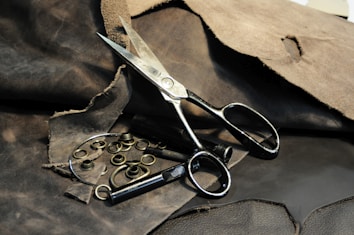 A pair of large, metal scissors placed on a rough, dark brown leather surface. Surrounding the scissors are several metal grommets and tools intended for leatherwork or crafting. The leather in the background has a worn texture, with some pieces appearing frayed or torn.
