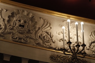 An intricately carved plaster frieze features a dramatic mask with open mouth, surrounded by ornate floral patterns. A candelabra with lit bulbs extends from the wall, casting warm light over the decorative elements.