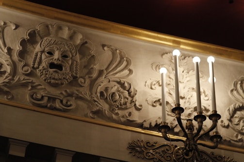 An intricately carved plaster frieze features a dramatic mask with open mouth, surrounded by ornate floral patterns. A candelabra with lit bulbs extends from the wall, casting warm light over the decorative elements.