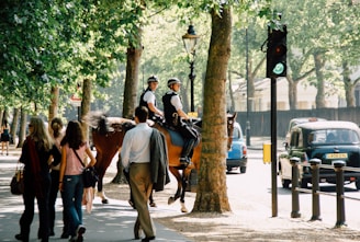 Two police officers on horseback are patrolling a tree-lined street as pedestrians pass by. Several people are walking in the opposite direction, some with shopping bags. A black cab is stopped at a traffic light, and a blue car is parked further down the street. The sunlight filters through the leaves, creating dappled shadows on the pavement.