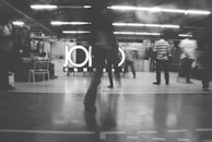 A black and white image of several people in motion inside a brightly lit industrial-style space. The background displays a large sign with the word 'CONCEPT' partially obscured by a person standing in the foreground. There are some objects like chairs and tables visible around the area.