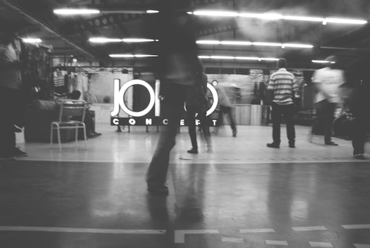 A black and white image of several people in motion inside a brightly lit industrial-style space. The background displays a large sign with the word 'CONCEPT' partially obscured by a person standing in the foreground. There are some objects like chairs and tables visible around the area.