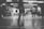 A black and white image of several people in motion inside a brightly lit industrial-style space. The background displays a large sign with the word 'CONCEPT' partially obscured by a person standing in the foreground. There are some objects like chairs and tables visible around the area.