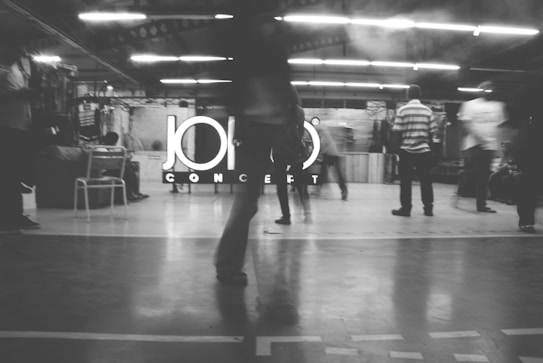 A black and white image of several people in motion inside a brightly lit industrial-style space. The background displays a large sign with the word 'CONCEPT' partially obscured by a person standing in the foreground. There are some objects like chairs and tables visible around the area.