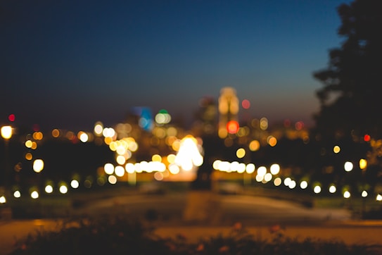 A cityscape at night with blurred lights and bokeh effect, featuring various colored lights scattered across the scene. The sky is dark with a subtle gradient transitioning from deep blue to black.