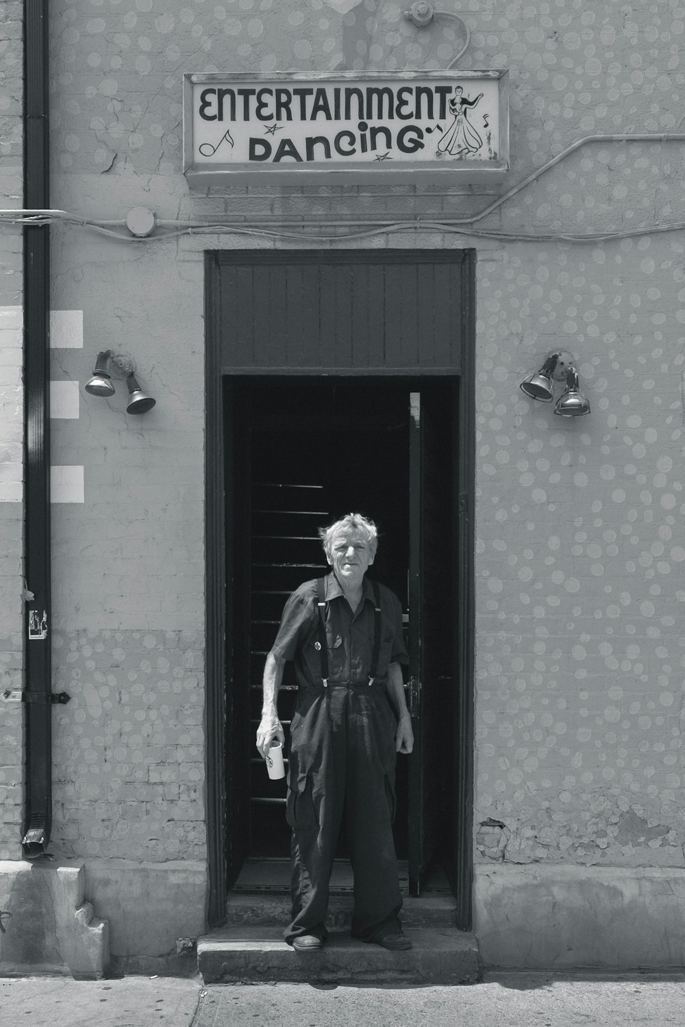 grayscale photo of man wearing collared tops and dress pants standing between concrete wall near Entertainment Dancing signage building