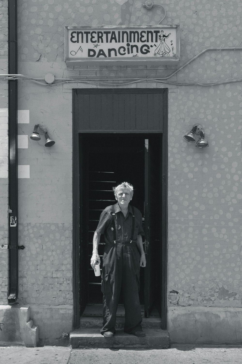 Foto en escala de grises de un hombre con blusas con cuello y pantalones de vestir de pie entre una pared de concreto cerca del edificio de señalización de Entertainment Dancing