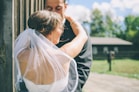 A bride in a white dress and veil embraces a groom dressed in a dark suit. The scene appears to be outdoors in a rural setting, with a wooden structure and a blurry view of trees and a building in the background.