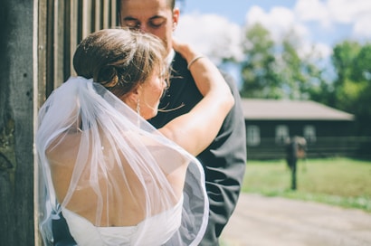 A bride in a white dress and veil embraces a groom dressed in a dark suit. The scene appears to be outdoors in a rural setting, with a wooden structure and a blurry view of trees and a building in the background.