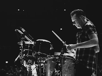 A drummer intensely playing an instrument, with a focused expression and a slight spotlight on the face, surrounded by a dark, ambient setting. The drum kit is well-lit, highlighting its metallic and polished surfaces.