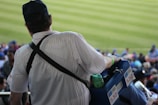 A person wearing a striped white shirt and a dark cap carries a blue cooler box with several beverage cans and bottles. They appear to be a vendor at a sporting event, with a strap slung over their shoulder. In the background, blurred spectators are seated, and a well-maintained green field is visible.