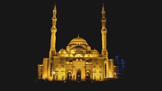 An illuminated mosque stands majestically against a dark night sky. The architecture features multiple domes and two tall minarets on either side, all lit with a warm golden hue. Ornate arches and geometric design elements enhance the building's facade, creating a sense of grandeur and serenity.
