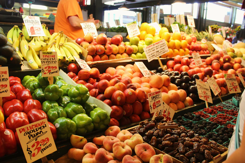 A farmers market produce stand piled with bananas, peppers, apples, peaches, and berries with hand-written price signs