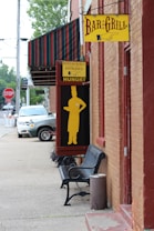 A street view featuring the side of a brick building with a yellow 'Bar and Grill' sign. Below, another sign indicates a restaurant entrance with a silhouette of a person. A black bench with a small trash bin is placed on the sidewalk, and a white vehicle is parked nearby. The awning of the building is striped in dark colors, and some greenery can be seen in the background.