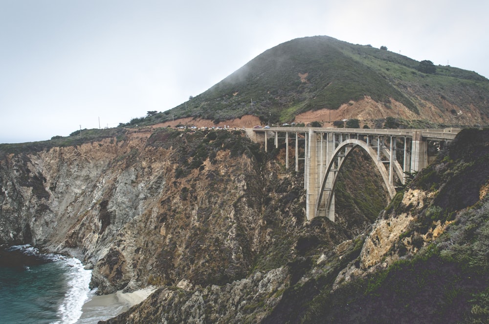 日中の水域近くの茶色と黒の崖の間の灰色のコンクリート橋