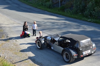 A classic car is parked on a rural road, with a woman and two children nearby. The woman is kneeling with a colorful backpack beside her, while one child stands wearing sunglasses and the other sits on the car's fender.