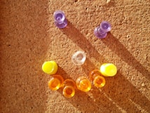 A corkboard is decorated with colorful push pins arranged to form a smiling face. Two purple push pins represent the eyes, while a clear push pin is used for the nose. The mouth is formed by alternating yellow and orange push pins.