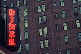 A neon sign displaying the word 'DINER' is attached to a brick building with numerous windows, creating a vintage or nostalgic atmosphere.