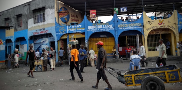 A bustling street scene with numerous people engaged in various activities outside a row of shops. The shops have vibrant signage in different colors and languages. Some individuals are walking, while others are interacting or sitting. A man pulls a cart in the foreground.