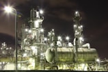 An industrial complex at night with numerous illuminated structures and pipes, giving off bright lights against the dark sky. The metallic surfaces of the machinery reflect the glow, creating a futuristic and bustling atmosphere.