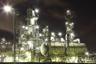 An industrial complex at night with numerous illuminated structures and pipes, giving off bright lights against the dark sky. The metallic surfaces of the machinery reflect the glow, creating a futuristic and bustling atmosphere.