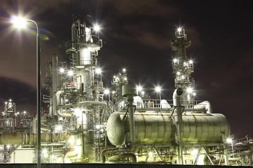 An industrial complex at night with numerous illuminated structures and pipes, giving off bright lights against the dark sky. The metallic surfaces of the machinery reflect the glow, creating a futuristic and bustling atmosphere.