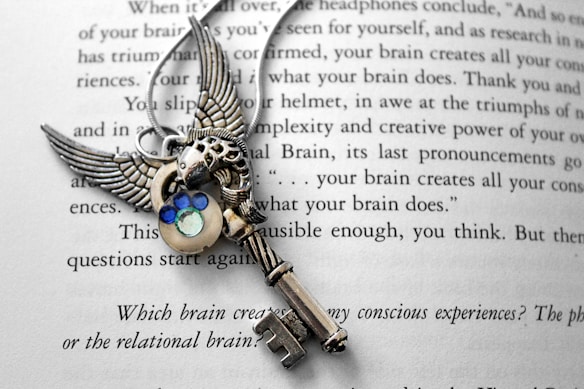 A silver ornate key with wings and decorative blue and green gemstones is placed on an open book. The page of the book contains text discussing consciousness and the brain.