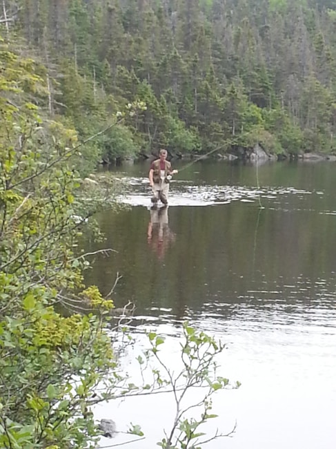 A person stands with waders in a shallow part of a lake, surrounded by dense coniferous forest. The individual appears to be fishing, holding a fishing rod while being partially submerged in water. The scene is calm and serene, with reflections of the trees on the water's surface.