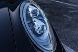 Close-up of a car's headlight featuring a sleek, modern design with intricate reflective components. The surface of the headlight showcases sharp lines and a glossy finish. The surrounding body of the car is dark and polished, contributing to a luxurious vibe.