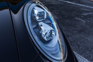 Close-up of a car's headlight featuring a sleek, modern design with intricate reflective components. The surface of the headlight showcases sharp lines and a glossy finish. The surrounding body of the car is dark and polished, contributing to a luxurious vibe.