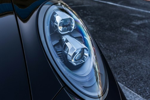Close-up of a car's headlight featuring a sleek, modern design with intricate reflective components. The surface of the headlight showcases sharp lines and a glossy finish. The surrounding body of the car is dark and polished, contributing to a luxurious vibe.