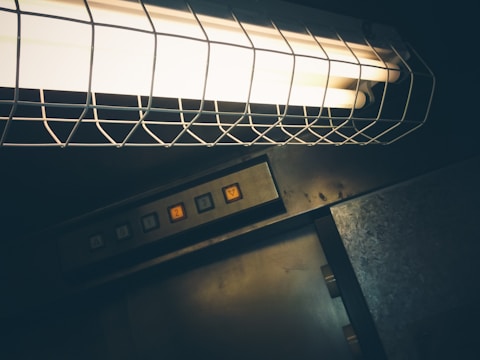 A dimly lit environment featuring a fluorescent light encased in a wire cage. Below the light, there is a metallic panel with elevator buttons labeled 1, 2, 3, and a triangular symbol for navigation.