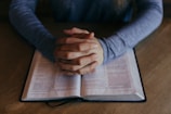 A person sitting at a wooden table with hands clasped together in a pose of reflection or prayer over an open book.