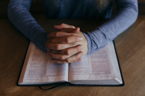 A person sitting at a wooden table with hands clasped together in a pose of reflection or prayer over an open book.