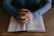 A person sitting at a wooden table with hands clasped together in a pose of reflection or prayer over an open book.