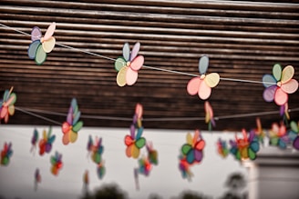 Colorful paper flowers are strung on thin wires under a roof of parallel wooden slats. Each flower consists of multiple pastel and bright-colored petals, creating a cheerful and lively ambiance. The background features blurred greenery.