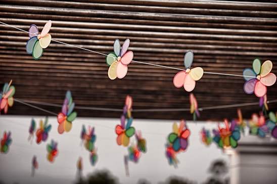 Colorful paper flowers are strung on thin wires under a roof of parallel wooden slats. Each flower consists of multiple pastel and bright-colored petals, creating a cheerful and lively ambiance. The background features blurred greenery.