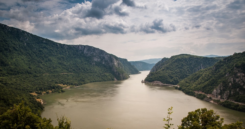 grey calm body of water between mountains and tall trees under white clouds at daytime