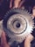 A close-up view of a metallic gear held by two hands. The gear features a central bearing surrounded by small, evenly spaced teeth. Slight wear and grime are visible, suggesting use in a mechanical environment. The hands holding the gear are slightly dirty, indicating manual labor or repair work.