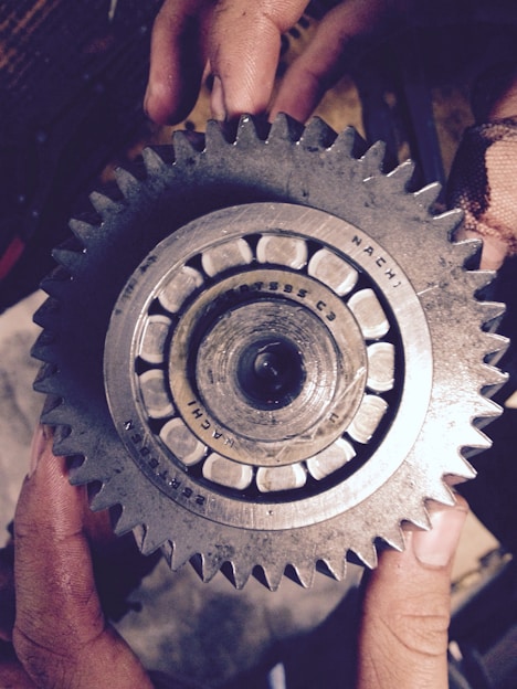 A close-up view of a metallic gear held by two hands. The gear features a central bearing surrounded by small, evenly spaced teeth. Slight wear and grime are visible, suggesting use in a mechanical environment. The hands holding the gear are slightly dirty, indicating manual labor or repair work.