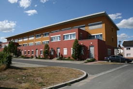 The image depicts a modern building with a combination of orange and red exteriors. It has multiple windows, a flat roof, and well-maintained greenery, including small trees and bushes, surrounding it. A dark-colored car is parked on the pavement close to the building. The sky is clear with scattered small white clouds.