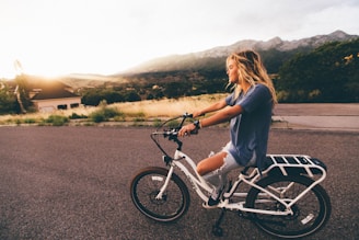 woman in blue t-shirt and distressed blue denim jeans riding white bicycle on grey road near mountains at daytime