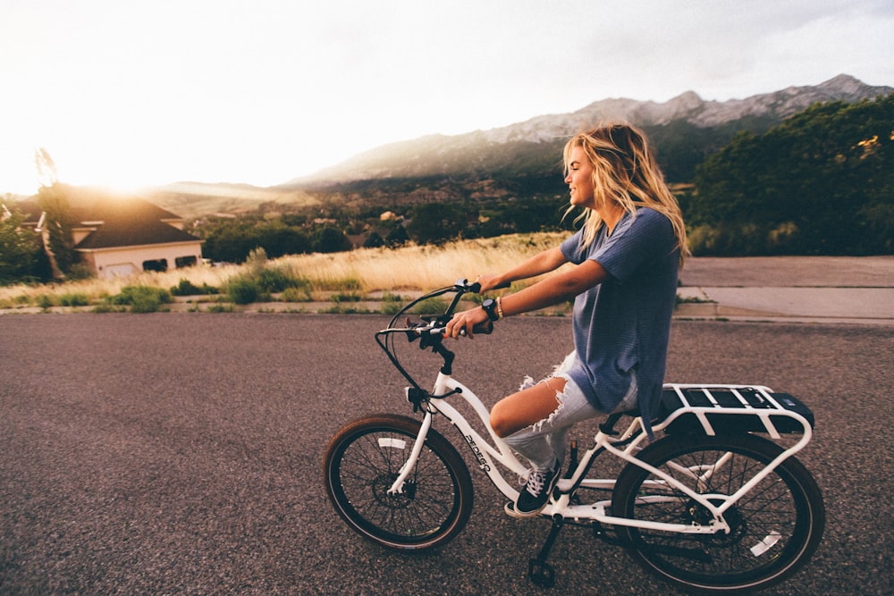 파란 티셔츠와 낡은 파란 데님 청바지를 입은 여자가 낮에 산 근처의 회색 도로에서 흰색 자전거를 타고 있습니다.