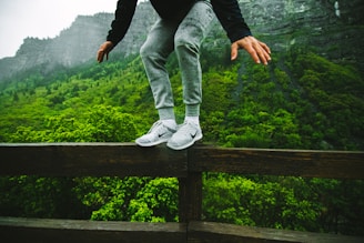 A person wearing gray sweatpants and white sneakers is balancing on a wooden railing with lush green forest and rocky mountains in the background. The scene conveys a sense of adventure and focus.