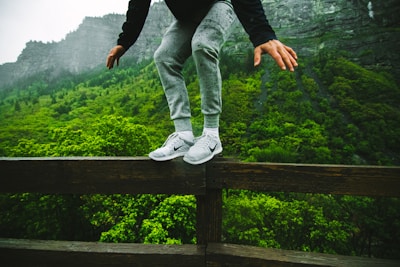 A person wearing gray sweatpants and white sneakers is balancing on a wooden railing with lush green forest and rocky mountains in the background. The scene conveys a sense of adventure and focus.