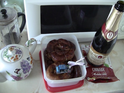 On a kitchen counter, there is a teapot with floral designs next to a French press. A plastic container filled with coiled sausages has an electronic component placed on top. A bottle of sparkling wine stands to the right of the container. A packet with labels in Cyrillic script is partially visible near the bottle. In the background, there is a white microwave oven.