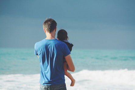 man wears blue crew-neck t-shirt holding toddler wears black hooded jacket near ocean under blue sky at daytime
