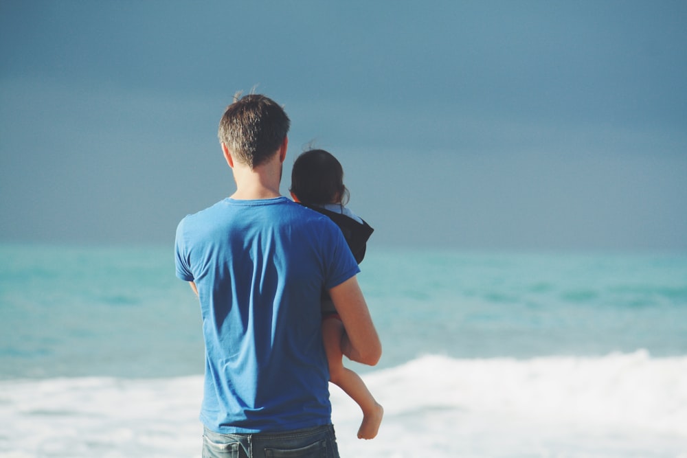 man wears blue crew-neck t-shirt holding toddler wears black hooded jacket near ocean under blue sky at daytime