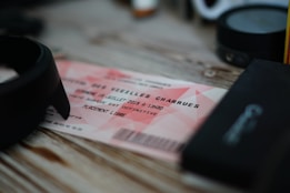 A concert ticket with the text 'Les Vieilles Charrues' printed on it, placed on a wooden surface. It is surrounded by various dark-colored objects, possibly electronic devices or accessories.