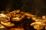 Several pieces of meat are being grilled on a barbecue, with smoke rising and a metallic tong in view, suggesting cooking in progress.